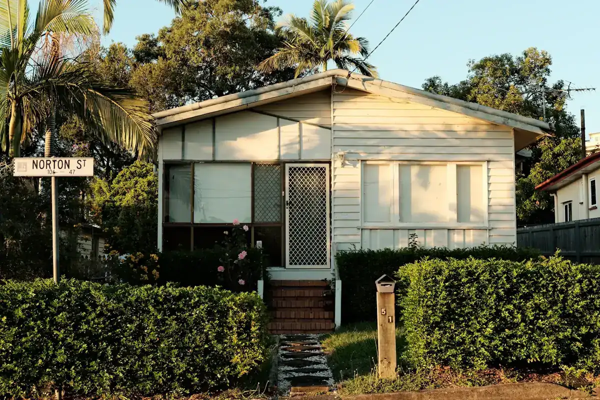 A timber Queenslander house behind a hedge on a suburban street